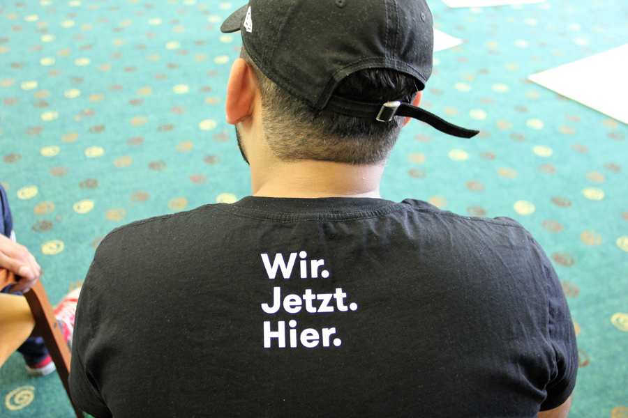 Jugendlicher mit Rücken zum Bild mit T-Shirt Aufschrift "Wir. Jetzt. Hier."