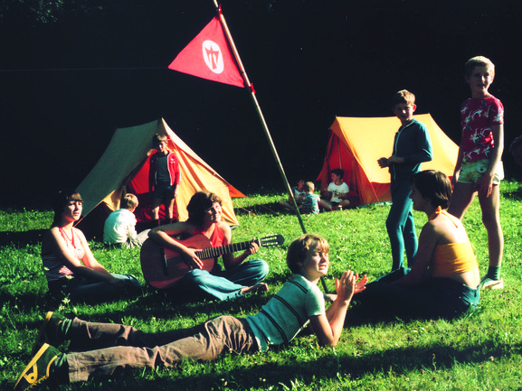 Jugendliche sitzen und liegen auf der Wiese, spielen Gitarre, dahinter Zelte